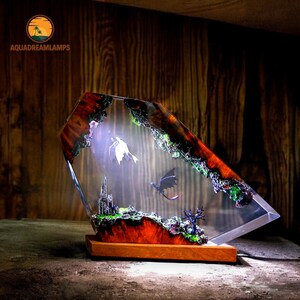 May include: A decorative lamp featuring a fantasy scene with a castle, dragons, and a glowing figure encased in a clear, geometric resin. The base is made of wood, and the lamp has a warm, inviting glow. The text "AQUADREAMLAMPS" is visible.