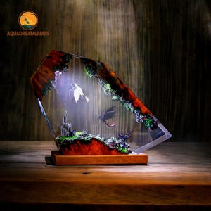 May include: A decorative resin lamp featuring a fantasy scene with two dragons, a castle, and a landscape. The lamp has a wooden base and a clear, angled resin body with embedded details. The lamp has the text "AQUADREAMLAMPS".