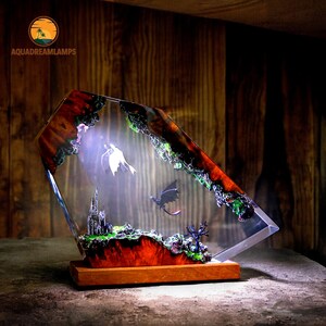 May include: A decorative lamp featuring a fantasy scene with two dragons, a castle, and a tree, encased in clear resin. The base is made of wood, and the lamp has a warm, inviting glow. The lamp is a unique piece of art.