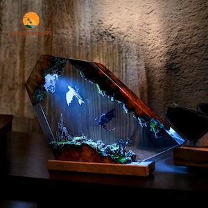 May include: A decorative resin lamp with a fantasy scene featuring two flying creatures, a castle, and a tree. The lamp has a wooden base and a geometric shape with a dark brown and green color scheme. The lamp is illuminated with a blue light.