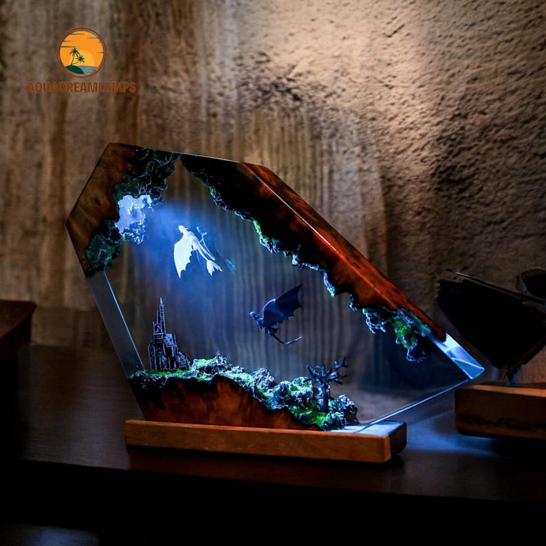 May include: A decorative resin lamp with a fantasy scene featuring two flying creatures, a castle, and a tree. The lamp has a wooden base and a geometric shape with a dark brown and green color scheme. The lamp is illuminated with a blue light.