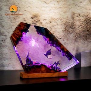 May include: A fantasy-themed lamp with a clear, angled resin body. Inside, a miniature castle, trees, and two dragon figures are illuminated with purple light. The lamp sits on a wooden base, creating a unique decorative piece.
