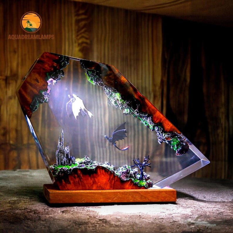 May include: A decorative resin lamp featuring a fantasy scene with dragons, a castle, and a white dragon. The lamp has a wooden base and a clear, angled resin body with a colorful, textured top. The lamp has the text "AQUADREAMLAMPS".