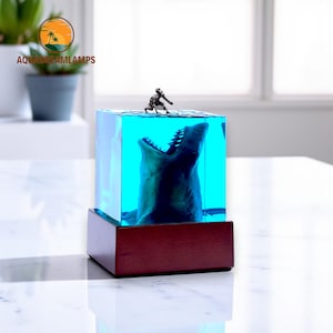 May include: A clear cube with a blue resin interior contains a shark head with an open mouth. A small figurine of a person is perched on the top of the cube, reaching towards the shark. The cube is on a wooden base with the text "AQUADREAMLAMPS" on the front.