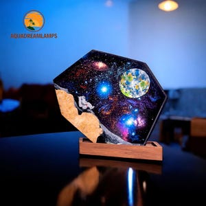 May include: A resin art piece depicting an astronaut sitting on the moon, with a galaxy background and a blue planet in the upper right corner. The piece is mounted on a wooden base.