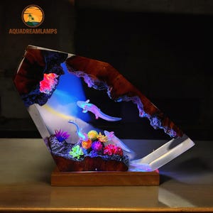 Puede incluir: Una lámpara de resina con un fondo azul, blanco y amarillo. La lámpara tiene una base de madera y una sección de resina transparente con dos axolotls, coral y otras plantas acuáticas. La lámpara está iluminada desde abajo, creando un brillo colorido.