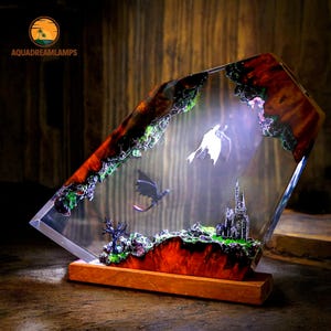 May include: A decorative resin lamp featuring a fantasy scene with two dragons, a castle, and a landscape. The lamp has a wooden base and the text "AQUADREAMLAMPS". The resin is a mix of red, brown, green, and clear. The lamp is lit from within.
