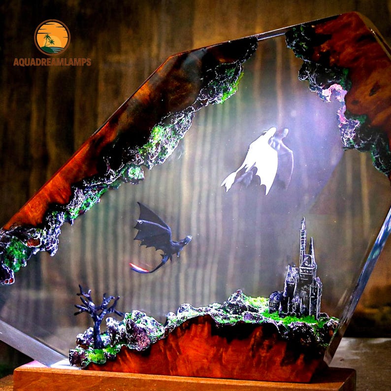 May include: A decorative lamp featuring a fantasy scene with dragons and a castle. The lamp has a clear, geometric shape with a wooden base. The edges are detailed with red, brown, and green accents, and the lamp includes the text "AQUADREAMLAMPS".