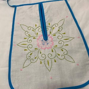 Può includere: Grembiule bianco con bordi e lacci blu. Presenta un ricamo floreale centrale rosa e verde, con accenti rosa. Il design è simmetrico e dettagliato, creando un oggetto decorativo e funzionale.