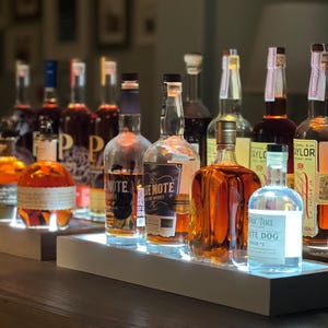May include: A collection of various glass liquor bottles arranged on two illuminated rectangular display stands. The bottles feature diverse labels, including Blue Note and Buffalo Trace White Dog Mash #1, with amber and clear liquids inside.