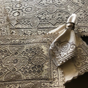 May include: An off-white linen table runner and matching placemat featuring intricate needlepoint lace borders with floral and geometric motifs. A folded napkin is secured with a metallic napkin ring, displaying a circular embroidered emblem.