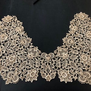 May include: An intricate, beige lace collar with a floral and scroll design. The delicate lace is arranged in a curved shape, showcasing detailed patterns and textures. The collar is likely for formal wear or a vintage-inspired garment.