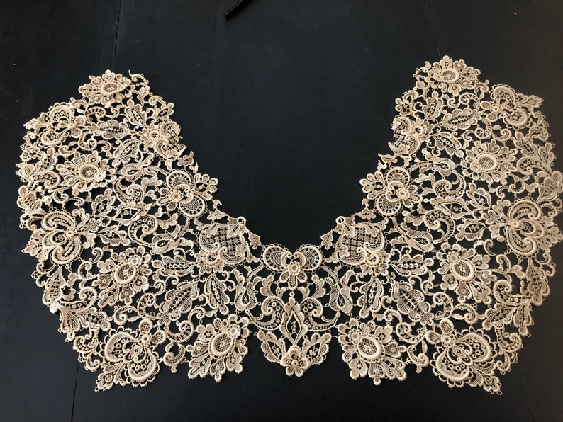May include: An intricate, beige lace collar with a floral and scroll design. The delicate lace is arranged in a curved shape, showcasing detailed patterns and textures. The collar is likely for formal wear or a vintage-inspired garment.