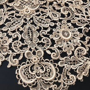 May include: Close-up of cream-colored lace fabric with intricate floral and scrollwork patterns. The delicate lace features detailed designs, including flowers and swirling motifs. The fabric is set against a dark background, highlighting the texture and detail of the lace.