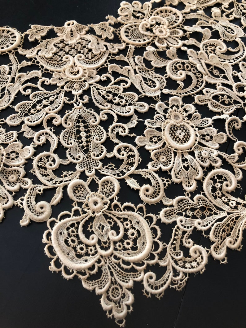May include: Close-up of cream-colored lace fabric with intricate floral and scrollwork patterns. The delicate lace features detailed designs, including flowers and swirling motifs. The fabric is set against a dark background, highlighting the texture and detail of the lace.