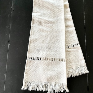 May include: A long, off-white runner featuring a textured, wavy woven pattern throughout. The fabric includes a horizontal band of openwork detailing near the bottom, finished with a fringed edge. The letters RB are embroidered in white thread.
