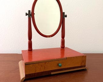May include: A tabletop vanity mirror featuring an oval glass pane framed in red wood, supported by two vertical red posts. The base is a rectangular wooden box with a single drawer, a small green knob, and a multi-toned wood grain finish.