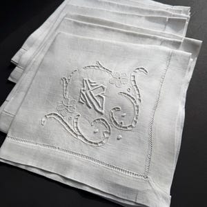 May include: A stack of white linen napkins. Each napkin features an embroidered floral design with an initial. The napkins have a decorative stitched border. The napkins are folded and stacked on top of each other.
