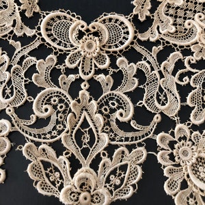 May include: Close-up of an off-white lace collar with intricate floral and scrollwork patterns. The delicate lace features a scalloped edge and is set against a dark background, highlighting the detailed craftsmanship.
