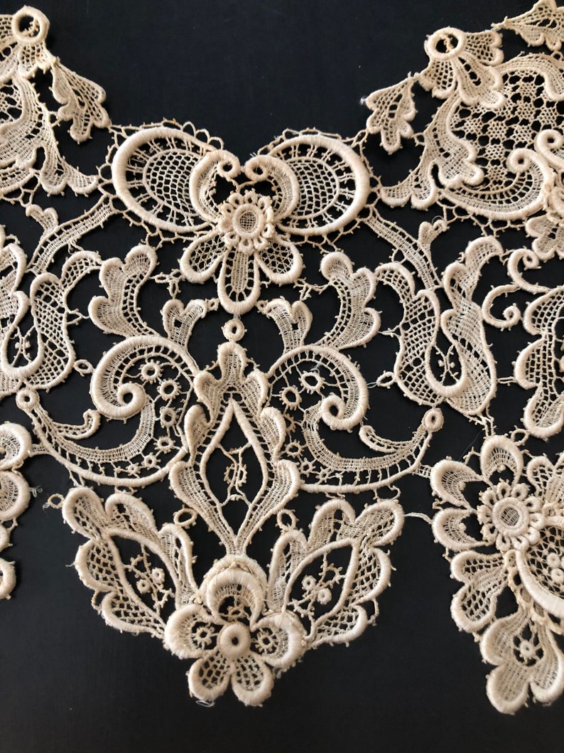 May include: Close-up of an off-white lace collar with intricate floral and scrollwork patterns. The delicate lace features a scalloped edge and is set against a dark background, highlighting the detailed craftsmanship.