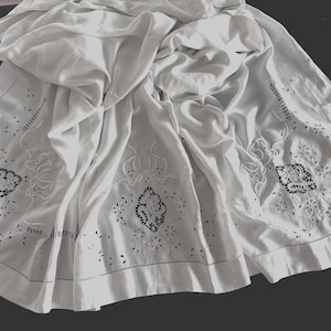 May include: White linen tablecloth with intricate cutwork and embroidered floral and geometric designs. The tablecloth is draped, showing the detailed edges and patterns. The cutwork includes small flower and leaf shapes, and the embroidery features a central medallion.