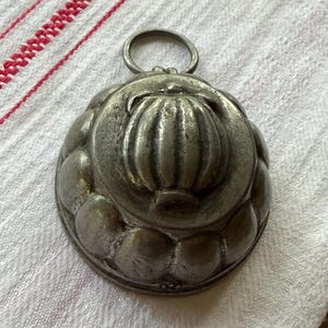 May include: A vintage metal jelly mold featuring a rounded, scalloped exterior design with a central embossed pumpkin or fruit motif and a small circular hanging loop attached to the top edge, showcasing a weathered, tarnished silver-toned finish.