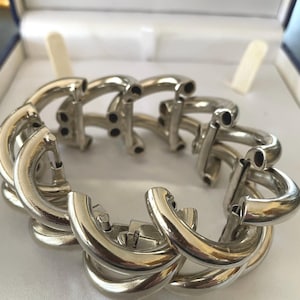 May include: A silver-toned metal bracelet composed of multiple interlocking curved tubular links. The design features a repeating pattern of polished, rounded segments that create a thick, chain-like structure with visible hollow ends on each individual link.