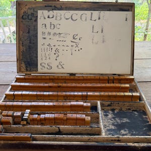 May include: A wooden box containing a collection of individual rubber stamps with wooden handles. The lid features a white paper sheet displaying various printed letters, numbers, and symbols including A, B, C, G, L, I, a, b, c, &, ?, and fractions.