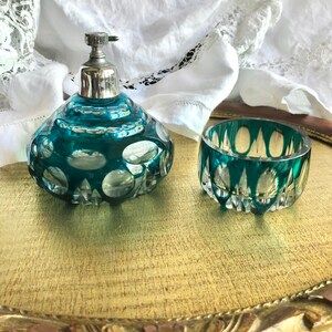 May include: A teal glass perfume bottle with a silver-toned metal pump mechanism and a matching teal glass open bowl, both featuring a cut-glass pattern of circular and triangular facets that catch the light, creating a clear and reflective appearance.