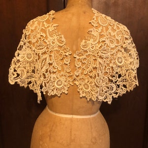 May include: Ivory lace shawl with intricate floral and scrollwork patterns. The delicate, openwork design is displayed on a mannequin, showcasing the shawl's shape and detail. A vintage-inspired accessory.