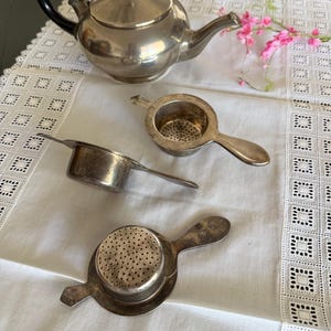 May include: A silver-toned metal teapot with a black handle and lid, accompanied by three matching silver-toned metal tea strainers. Each strainer features a circular bowl with a perforated base and a single handle, arranged on a white fabric surface.