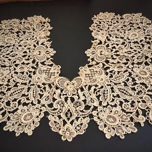 May include: Cream-colored lace collar with intricate floral and scrollwork patterns. The delicate, openwork design is symmetrical, with a V-neck shape. The lace is laid flat on a dark surface, showcasing its detailed craftsmanship.
