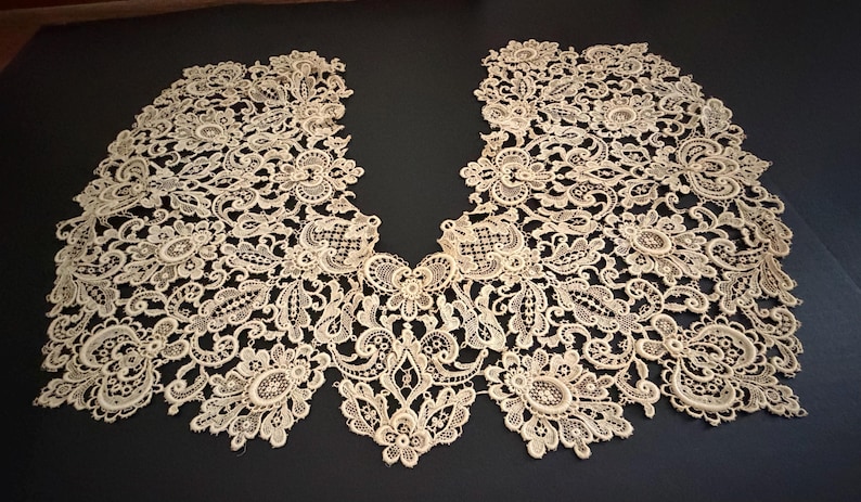 May include: Cream-colored lace collar with intricate floral and scrollwork patterns. The delicate, openwork design is symmetrical, with a V-neck shape. The lace is laid flat on a dark surface, showcasing its detailed craftsmanship.
