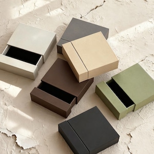 May include: A collection of square, sliding-drawer boxes in shades of beige, grey, brown, green, and black. Some boxes are open, showing a black interior. The boxes are displayed on a textured, light-coloured surface.