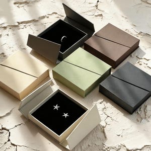 Può includere: Un assortimento di scatole portagioie in vari colori, tra cui beige, marrone, verde e nero. Alcune scatole sono aperte, rivelando un anello e orecchini a forma di stella su un interno in velluto nero. Le scatole sono disposte su una superficie strutturata.