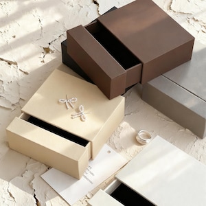 Puede incluir: Una variedad de joyeros en beige, marrón y gris. Las cajas están abiertas, revelando interiores de terciopelo negro. Se ven un par de pendientes de libélula plateados y un anillo plateado. Las cajas están dispuestas sobre una superficie texturizada.
