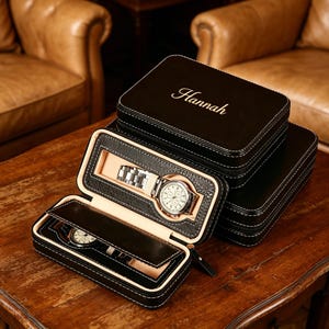 Puede incluir: Estuches de reloj de cuero negro con costuras blancas. Un estuche está abierto, mostrando un reloj plateado con esfera blanca. El nombre "Hannah" está grabado en la parte superior. Los estuches están sobre una superficie de madera.