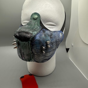 May include: A blue and green leather muzzle mask featuring a black nose section, silver-toned metal spikes on the sides, and a separate detachable bright red tongue piece. The mask is displayed on a white mannequin head with a black strap.