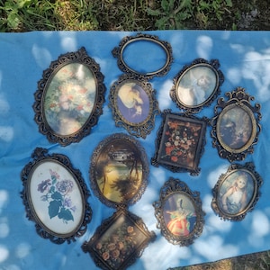 Puede incluir: Una colección de marcos de fotos antiguos de varias formas y tamaños, incluyendo ovalados, redondos y rectangulares. Los marcos son ornamentados, con un acabado bronce oscuro, y contienen obras de arte y retratos vintage. Los marcos están dispuestos sobre una superficie azul.