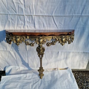 Può includere: Una mensola antica da parete con piano in legno scuro e ornati elementi in metallo color ottone. La mensola ha un bordo anteriore curvo e dettagli decorativi floreali e a volute. L'unica gamba di supporto è anch'essa color ottone.