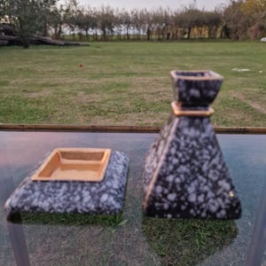 Può includere: Un set di due pezzi decorativi in ceramica. Uno è un posacenere quadrato con interno dorato. L'altro è un vaso a forma di piramide con una fascia dorata. Entrambi i pezzi hanno un motivo maculato bianco e nero.