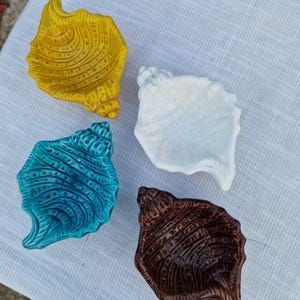 Könnte beinhalten: Vier keramische Muschelschalen in Gelb, Weiß, Türkis und Braun. Jede Schale hat eine strukturierte Oberfläche mit detaillierten Rillen, die einer natürlichen Muschel ähneln. Die Schalen sind auf einer weißen Stoffoberfläche angeordnet.