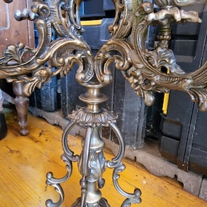 May include: Ornate, antique brass candelabra with intricate scrollwork and floral details. The candelabra has multiple arms and a detailed base, showcasing a vintage aesthetic. The finish is a warm, aged brass color, adding to its classic appeal.
