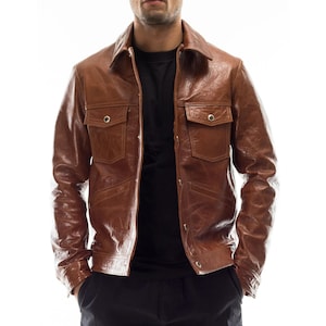 May include: A brown leather jacket with two chest pockets and a collared neckline. The jacket has a button-up front and a slightly distressed appearance. The model is wearing a black shirt underneath.