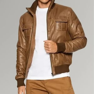 May include: A brown leather jacket with a zip-up front, two chest pockets, and a ribbed collar and cuffs. The jacket is worn over a white t-shirt and paired with brown pants. The jacket's color is a warm, medium brown.