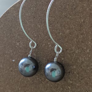 May include: A pair of dangle earrings featuring dark gray, round pearls suspended from curved, silver-toned wires. The earrings are displayed against a neutral background, highlighting their simple elegance and reflective surfaces.