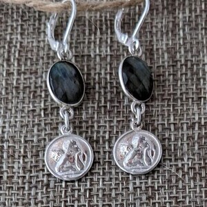 Peut inclure: Boucles d'oreilles pendantes en argent avec des pierres ovales gris fonc&eacute; et de petits m&eacute;daillons ronds repr&eacute;sentant une figure classique. Les boucles d'oreilles sont suspendues &agrave; des fermoirs &agrave; levier en argent.