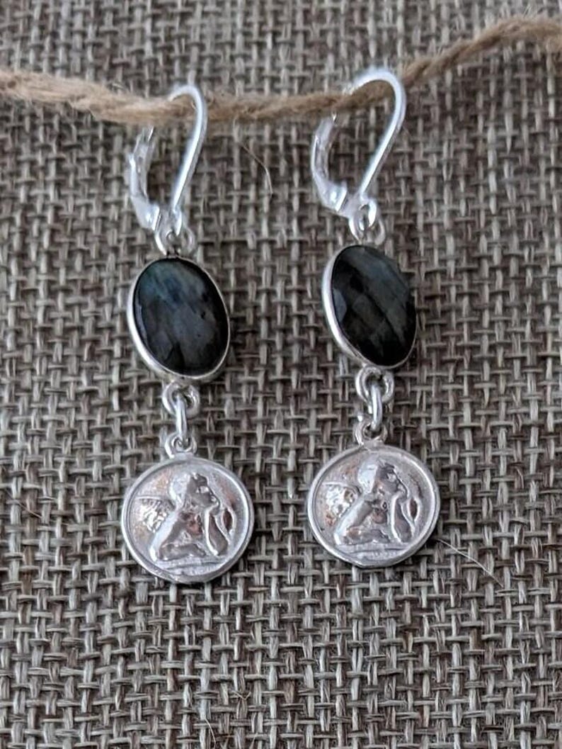 Peut inclure: Boucles d'oreilles pendantes en argent avec des pierres ovales gris fonc&eacute; et de petits m&eacute;daillons ronds repr&eacute;sentant une figure classique. Les boucles d'oreilles sont suspendues &agrave; des fermoirs &agrave; levier en argent.