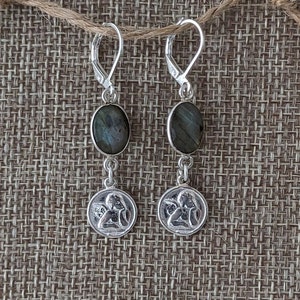 Peut inclure: Paire de boucles d'oreilles en argent avec des pierres ovales gris-vert et de petits pendentifs ronds repr&eacute;sentant un ange. Les boucles d'oreilles sont suspendues &agrave; des fermoirs &agrave; levier en argent.