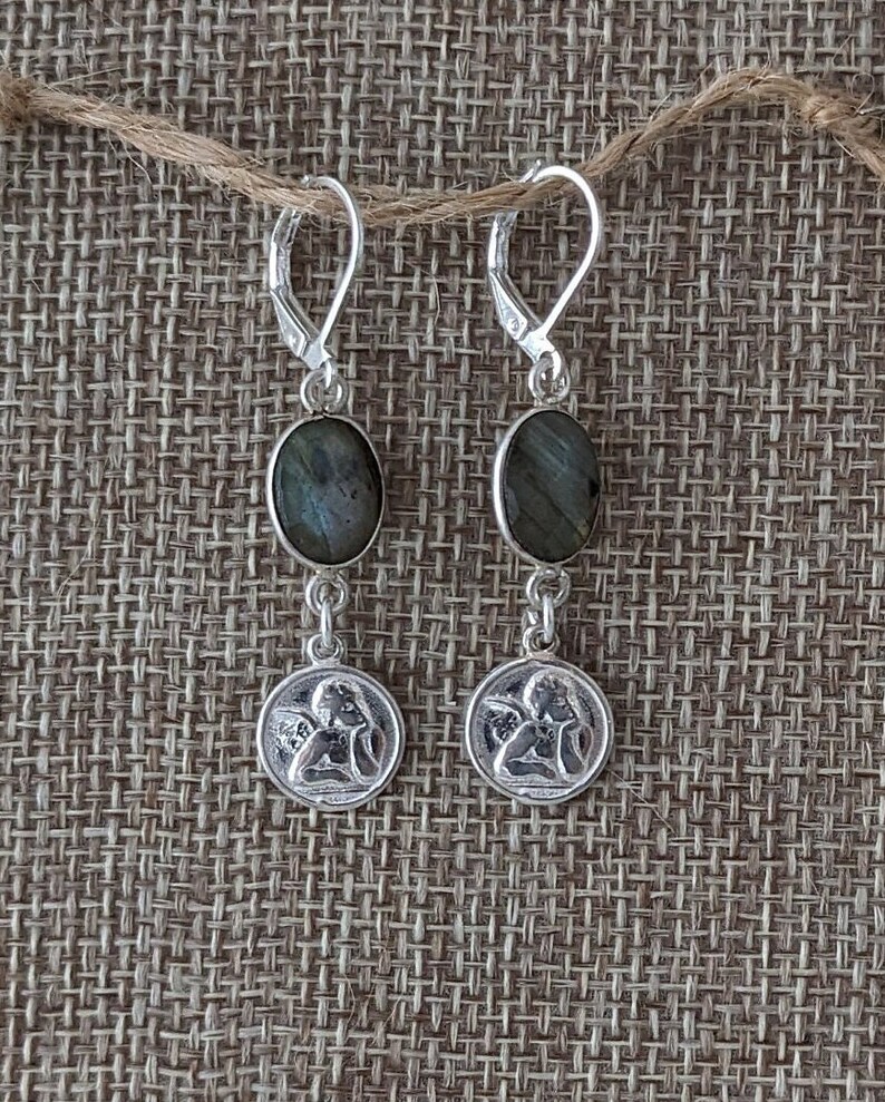 Peut inclure: Paire de boucles d'oreilles en argent avec des pierres ovales gris-vert et de petits pendentifs ronds repr&eacute;sentant un ange. Les boucles d'oreilles sont suspendues &agrave; des fermoirs &agrave; levier en argent.